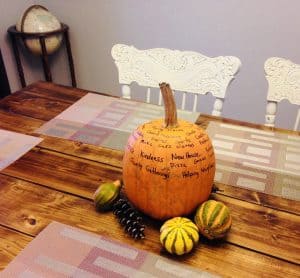 Photo of a "Thankful Pumpkin" sitting on a table. Words have been written on the pumpkin to reflect what the family is thankful for, such as "New House", "Kindness" and "Pizza".